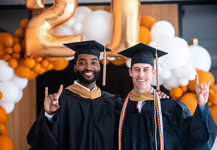 Two graduates give hook em horns in front of balloons