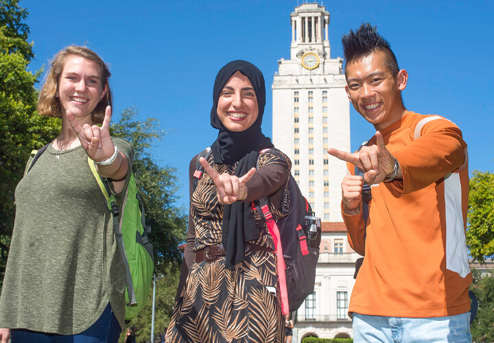 Three students in front of University of Texas tower giving hook em horns