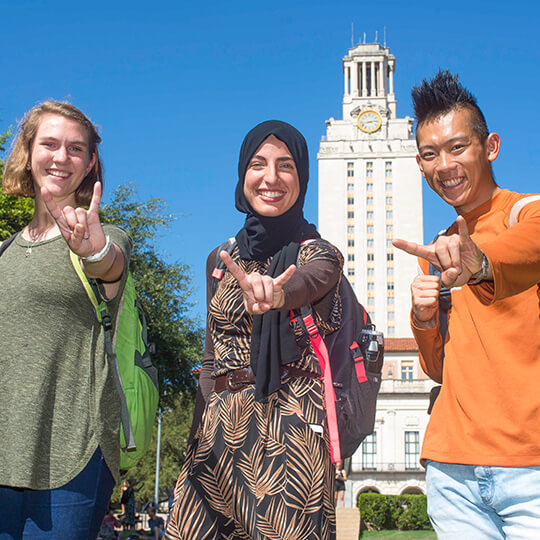 Three students in front of University of Texas tower giving hook em horns